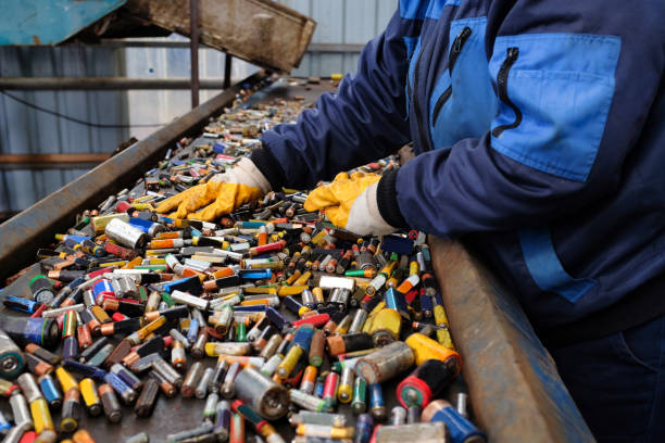 12 Inside a recycling facility, a worker in a blue jacket and yellow gloves is handling numerous used batteries on a metal conveyor, preparing them for the recycling process.